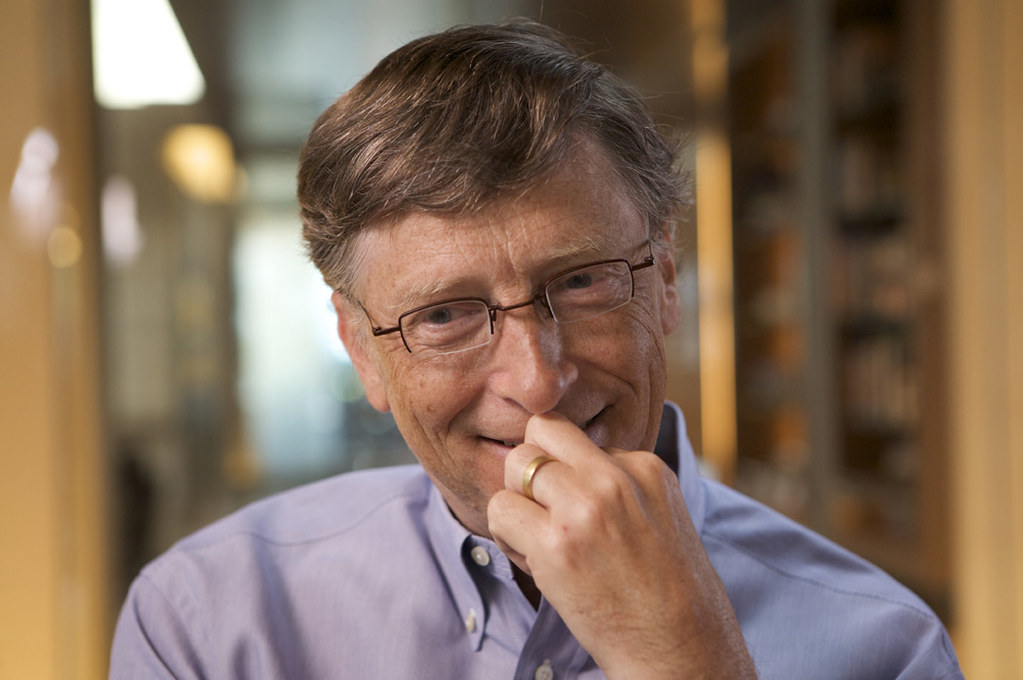 Bill gates grins with his hand near his mouth while wearing a light blue dress shirt