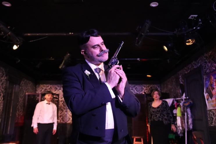 photo of an actor onstage wearing a wig and mustache and holding an antigue gun