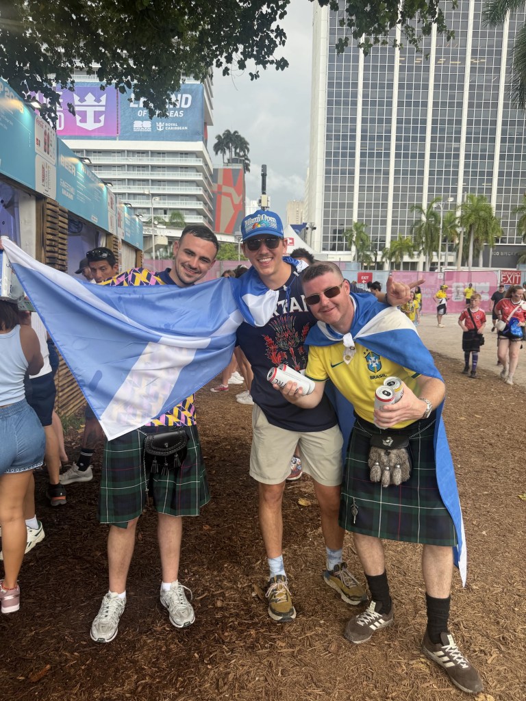 photo of three Scotland fans draped in their flag and sporting kilts at an outdoor festival