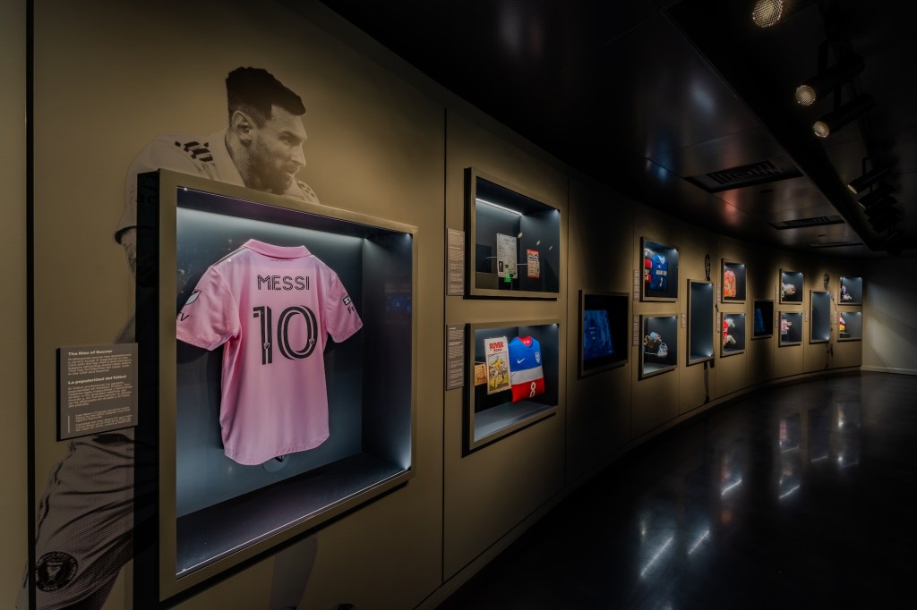 Photo of items on display inside a museum. On the left side of the wall is an image of soccer player Lionel Messi with his tee on display