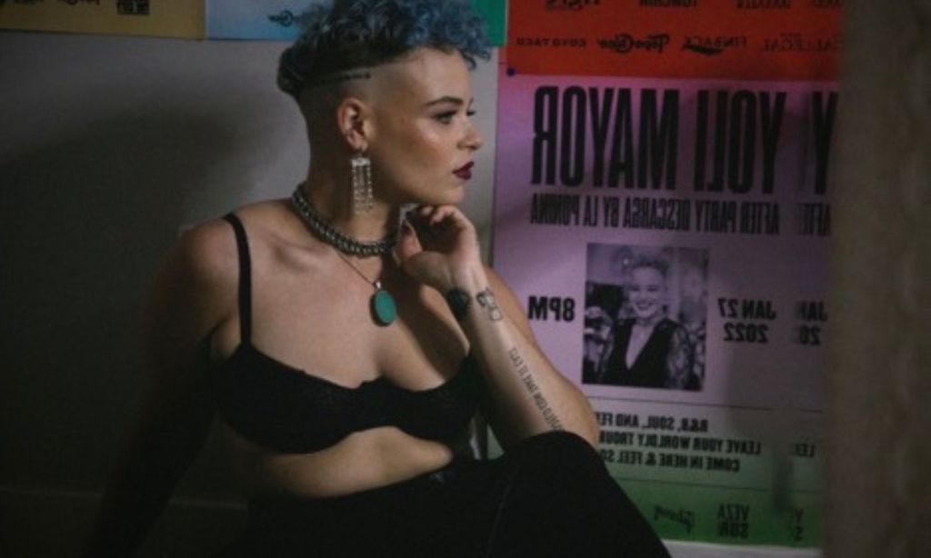Photo of a woman with short blue hair looking to the side wearing a black bra.