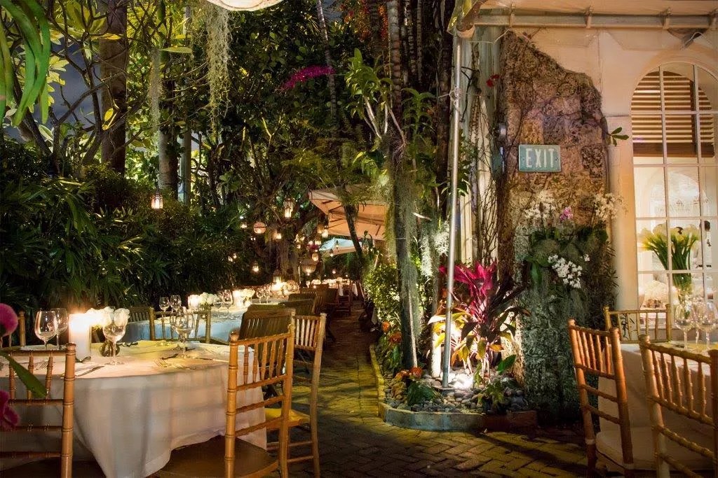 Photo of an outdoor restaurant with tables with tablecloths, candles, and place settings. Plants and trees surround the tables. The facade of the restaurant can be seen on the right, with windows peeking into the indoor dining room