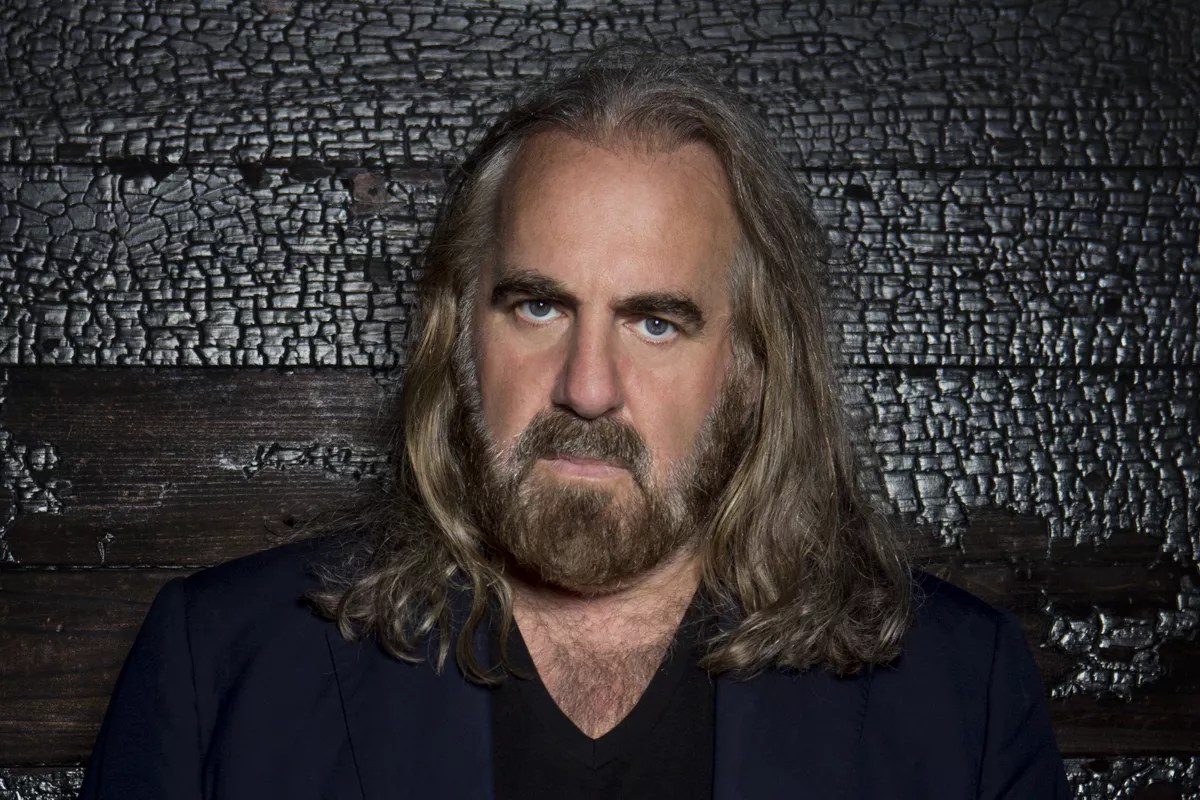 Photo of a man with long hair and a mustache standing in front of a wooden wall.