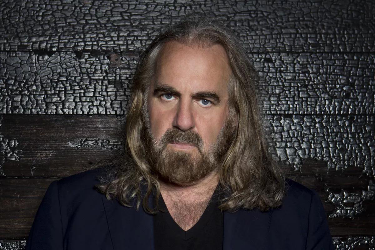 Photo of a man with long hair and a mustache standing in front of a wooden wall.