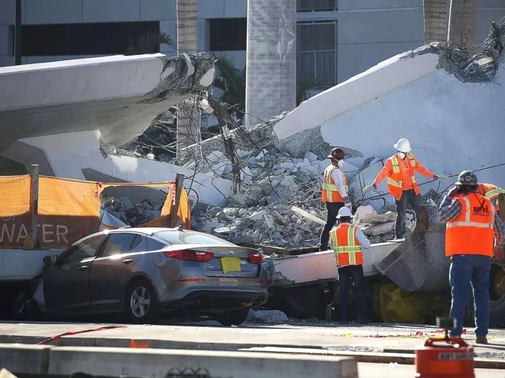 Construction workers in bright orange vests examine the debris-ridden site of the fatal Florida International University (FIU) bridge collapse. A gray Kia appears to be crushed beneath a large slab of concrete and mangled cables.