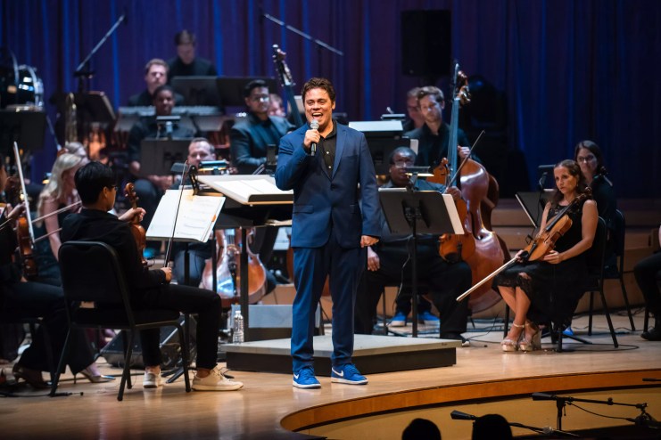 Nu Deco Ensemble composer Sam Hyken speaking on stage with the orchestra behind him