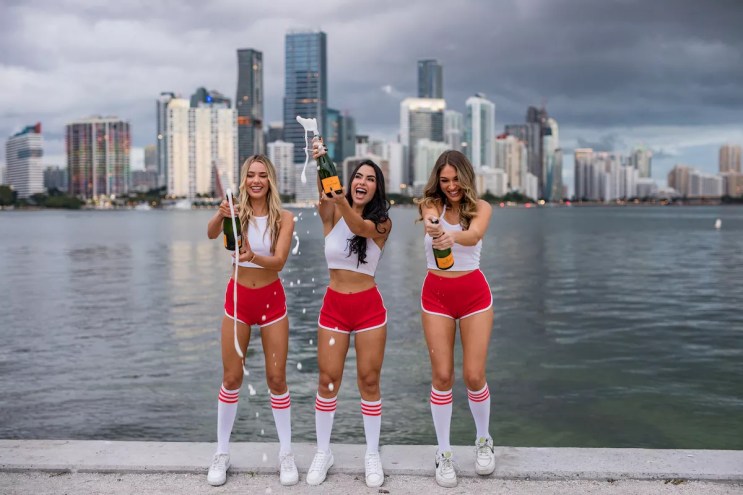 Three women in red shorts popping bottles of Champagne.