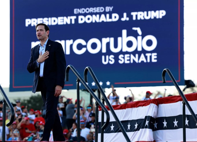 Color photo of Florida Republican Sen. Marco Rubio arriving at a political rally in Miami-Dade in the lead-up to the 2022 midterm elections. Rubio, wearing a blue suit, is shown in profile as he strides across the outdoor stage with his right hand covering his heart.