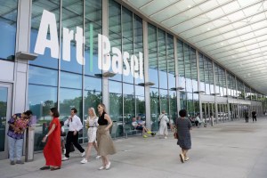 photo of people walking in front of the glass windows of a convention center with a sign on it reading, "Art Basel"