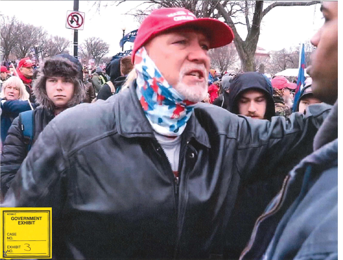 photo of a middle aged man wearing a red MAGA cap, multicolored gaiter, and black leather jacket, gesticulating in a crowd outside the US Capitol