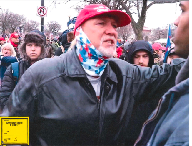 photo of a middle aged man wearing a red MAGA cap, multicolored gaiter, and black leather jacket, gesticulating in a crowd outside the US Capitol