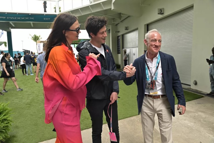 A woman in a pink suit and a man with brown hair, both smiling, stand with an elderly man wearing a lanyard