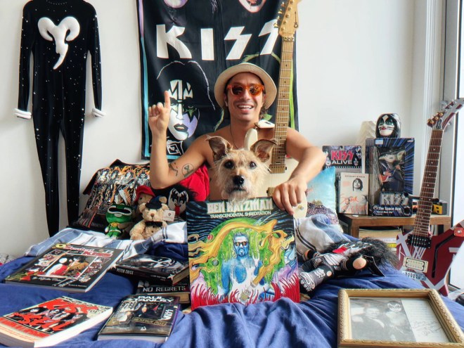 Musician Ben Katzman sits on his bed holding a copy of his album, Transcendental Shredidation, with a Kiss poster hanging on his wall in the background