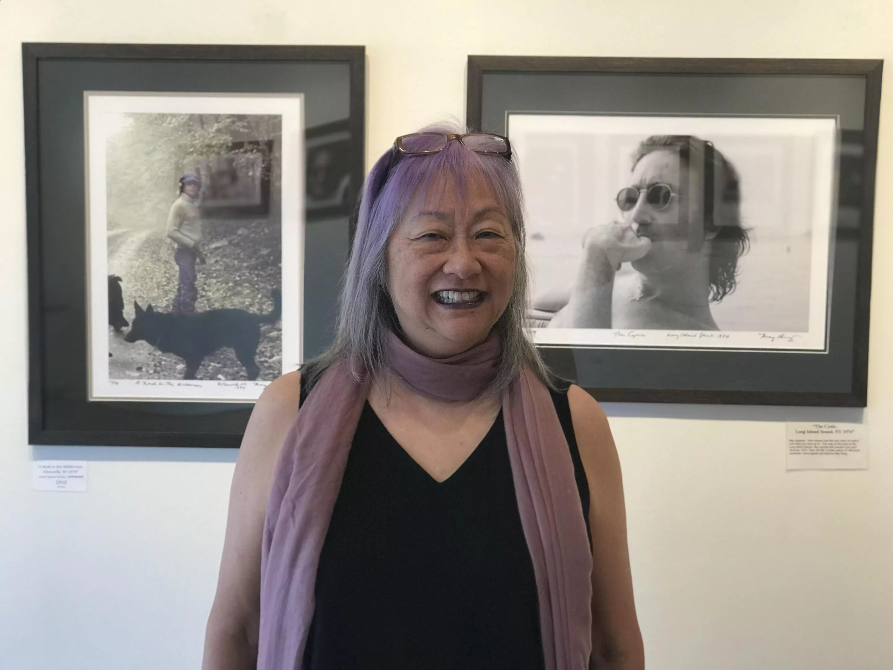 color photo of a smiling gray-haired woman in an art gallery with framed photos of John Lennon behind her