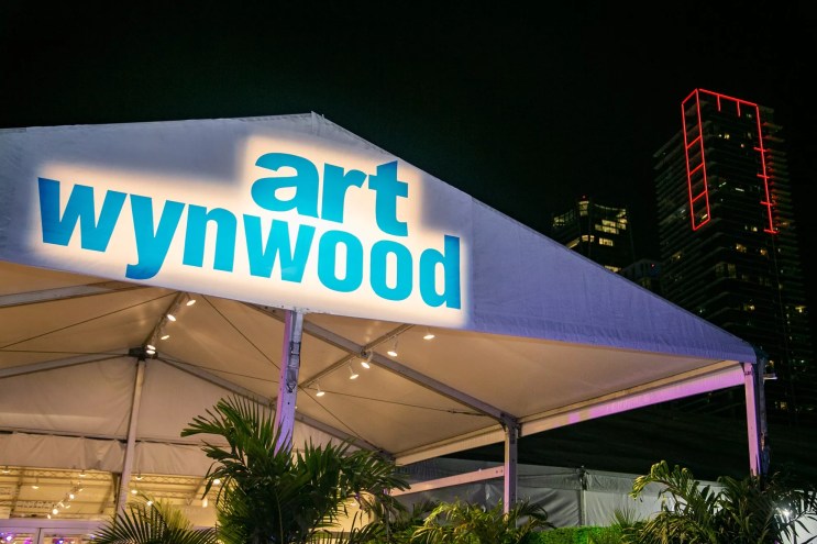 The words "Art Wynwood" spotlighted on the tent with the downtown Miami skyline in the background