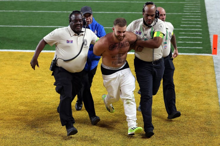 A shirtless Super Bowl streaker smiles as security guards haul him off the Allegiant Stadium turf.
