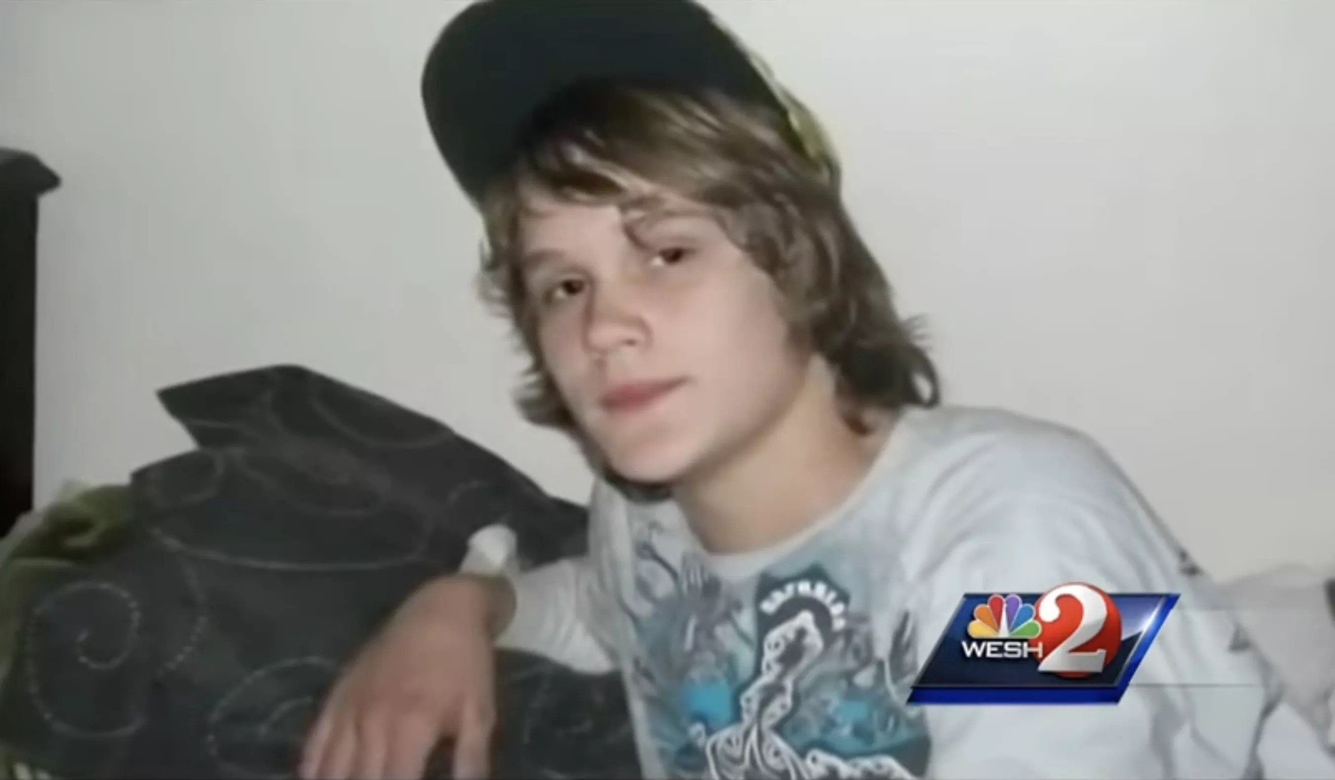 color snapshot of a young boy with shaggy shoulder-length brown hair, wearing a t-shirt and a black ball cap perched high on his head, looking into the camera