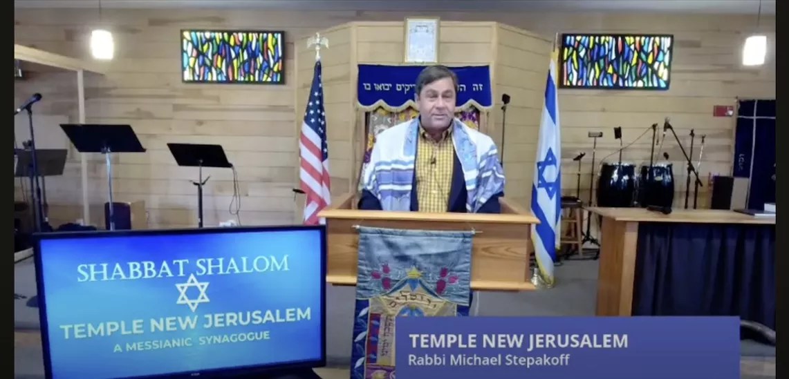 Michael Gary Stepakoff leads a shabbat service at his synagogue, Temple New Jerusalem. The 56-year-old is standing at a lectern before an Israeli and American flag and wearing a prayer shawl.