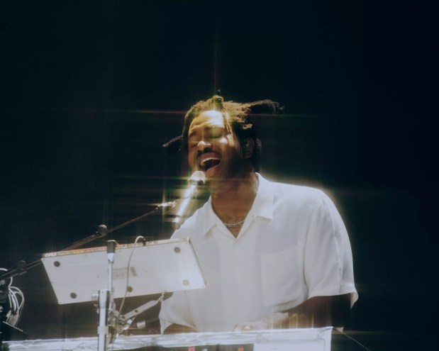 color photo of a white-shirted, dreadlocked Sampha singing into a microphone against a black background