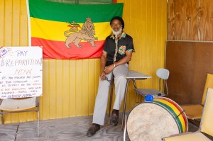Ras Iroy standing in front of the Rastafarian flag and a protest sign