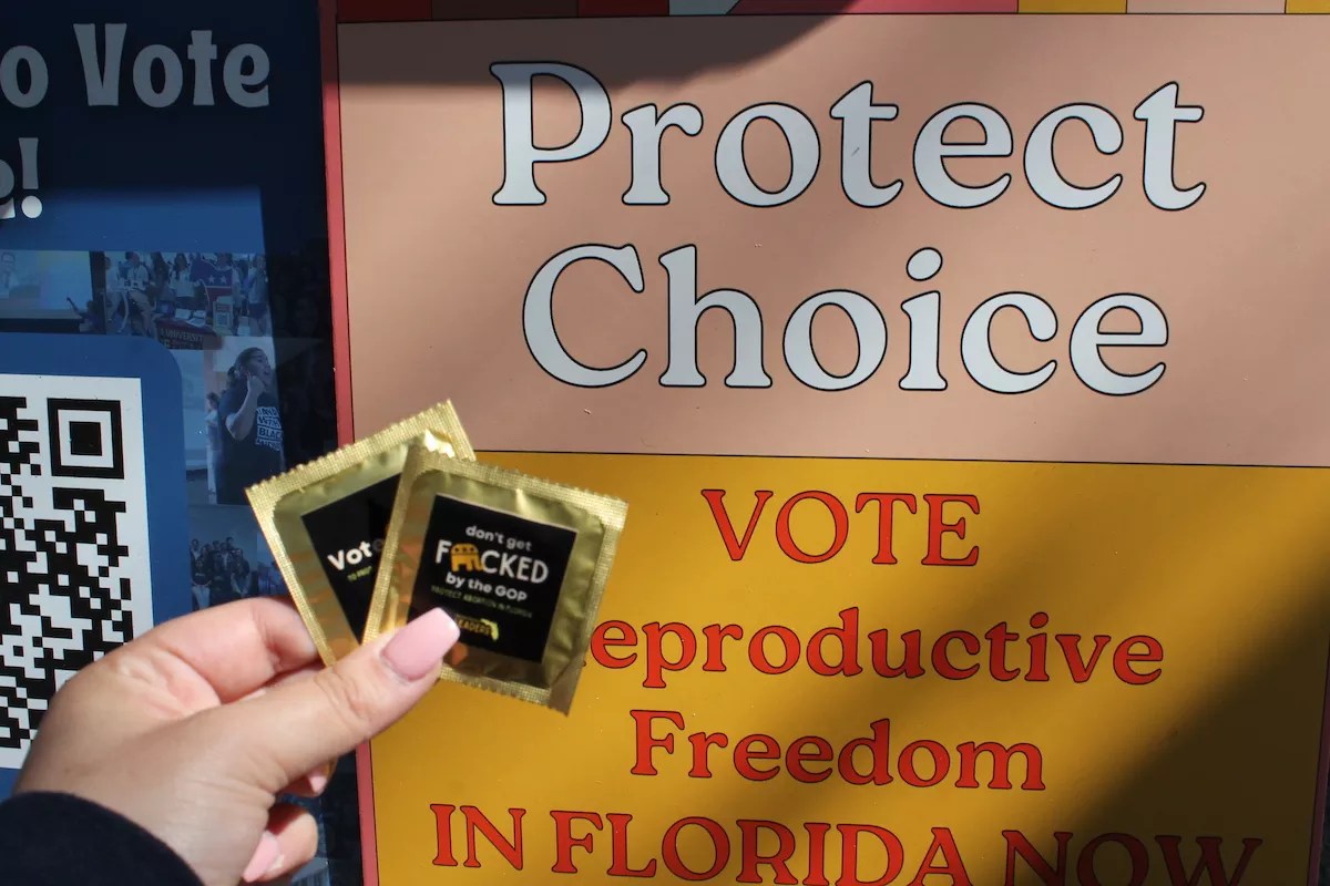 A hand holds two black and gold condoms with the words “Don’t Get Fucked by the GOP. Protect Abortion in Florida" printed on the front, in front of a yellow and orange sign that reads "Protect Choice, Vote Reproductive Freedom in Florida Now."