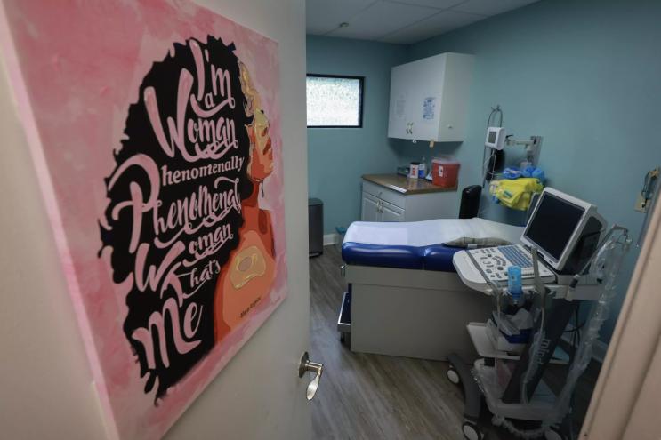 A door with a pink poster is propped open, leading to an examination room in an abortion clinic