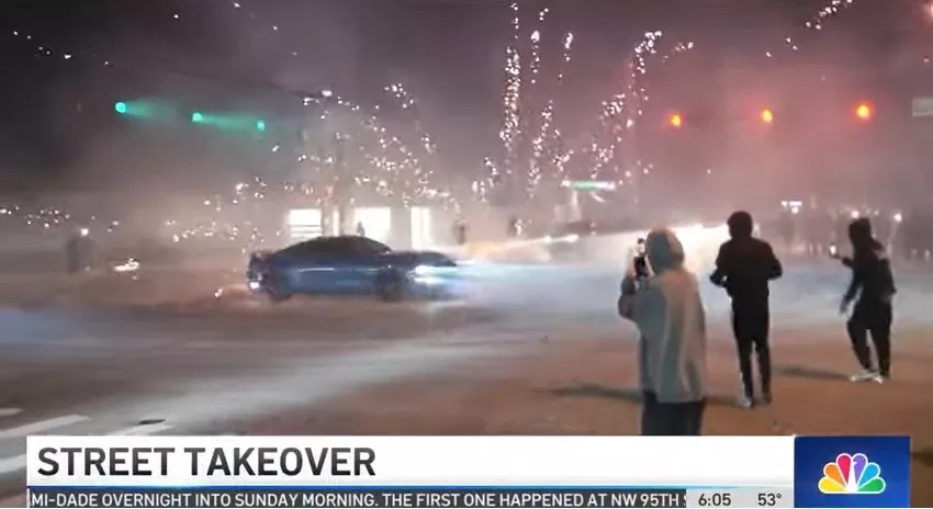 A car spins around an intersection during a street takeover as fireworks explode in the background
