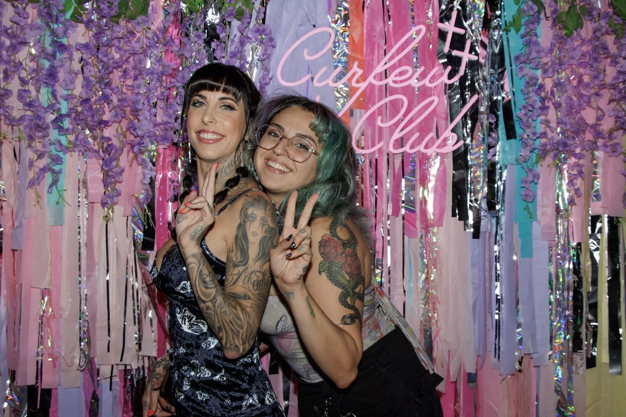 Two women pose in front of a Curfew Club backdrop