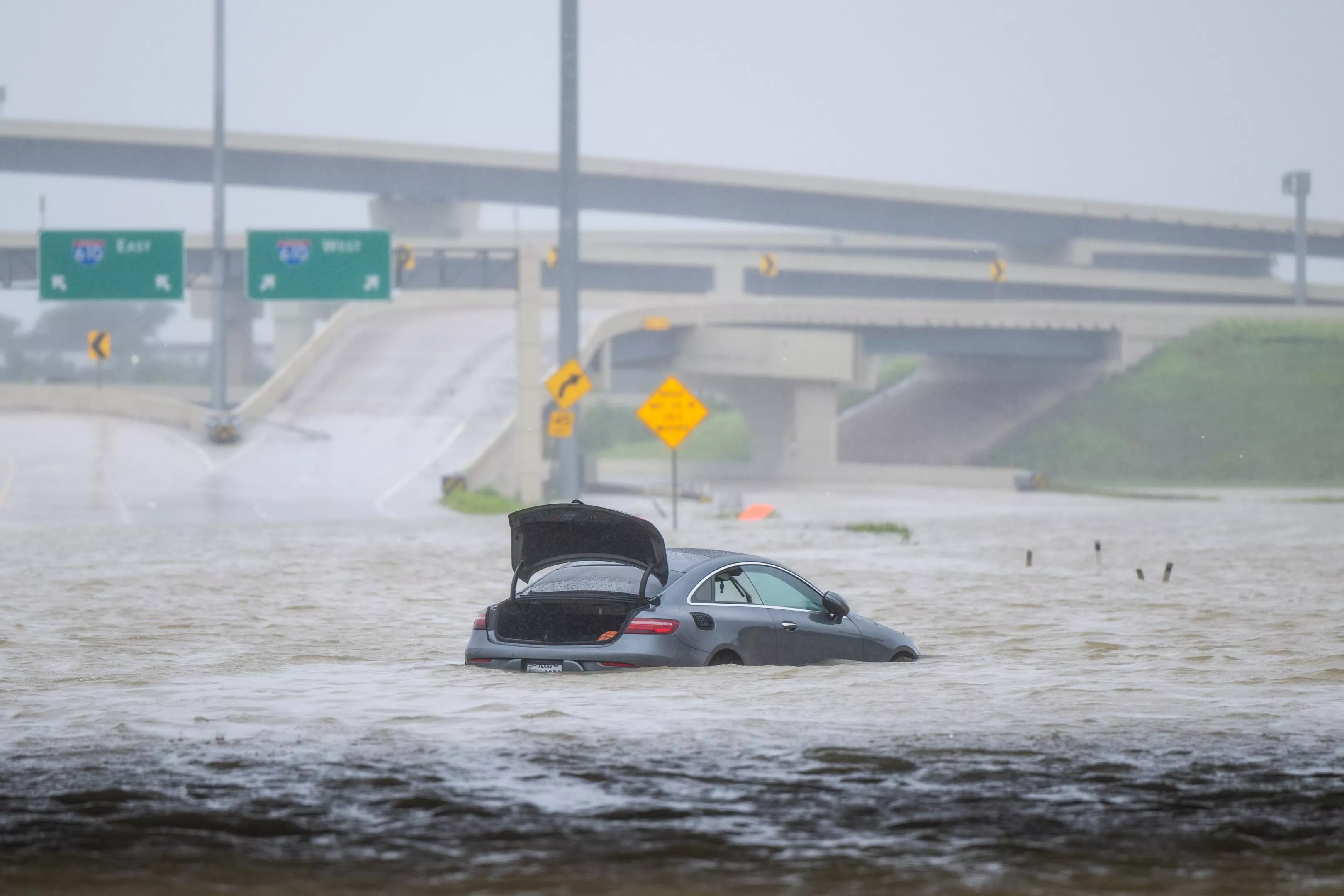 A vehicle with its trunk open in the middle of deep floodwaters on a highway