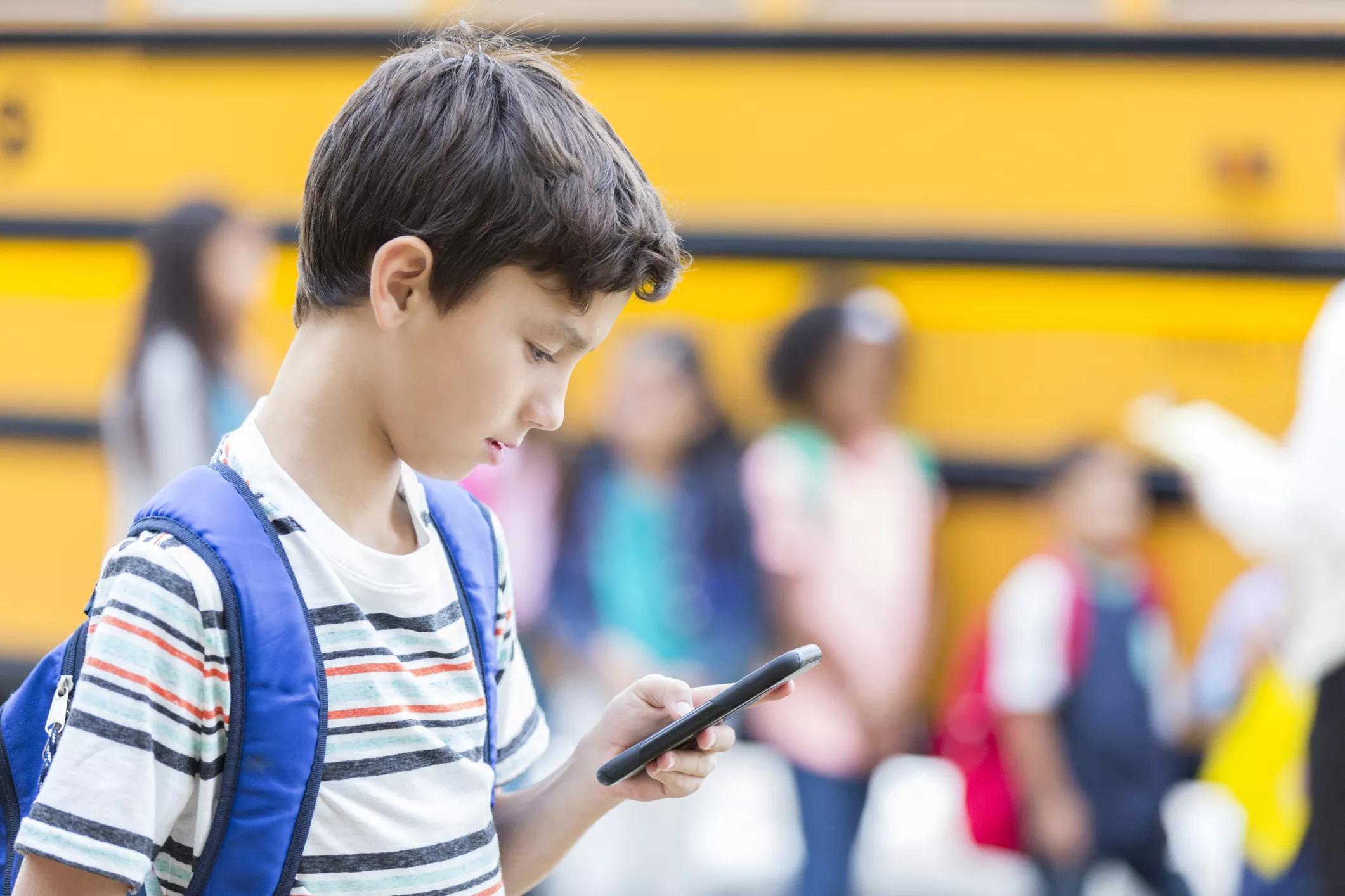 An elementary-school age boy with a blue backpack looks down at his cellphone in front of a school bus