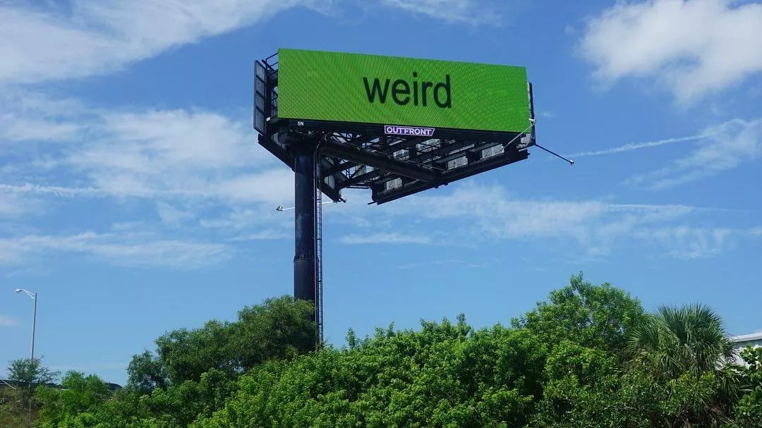 billboard with the word "weird" spelled out in lowercase sans serif letters against a bright green background, set against a verdant South Florida landscape on a bright sunny day