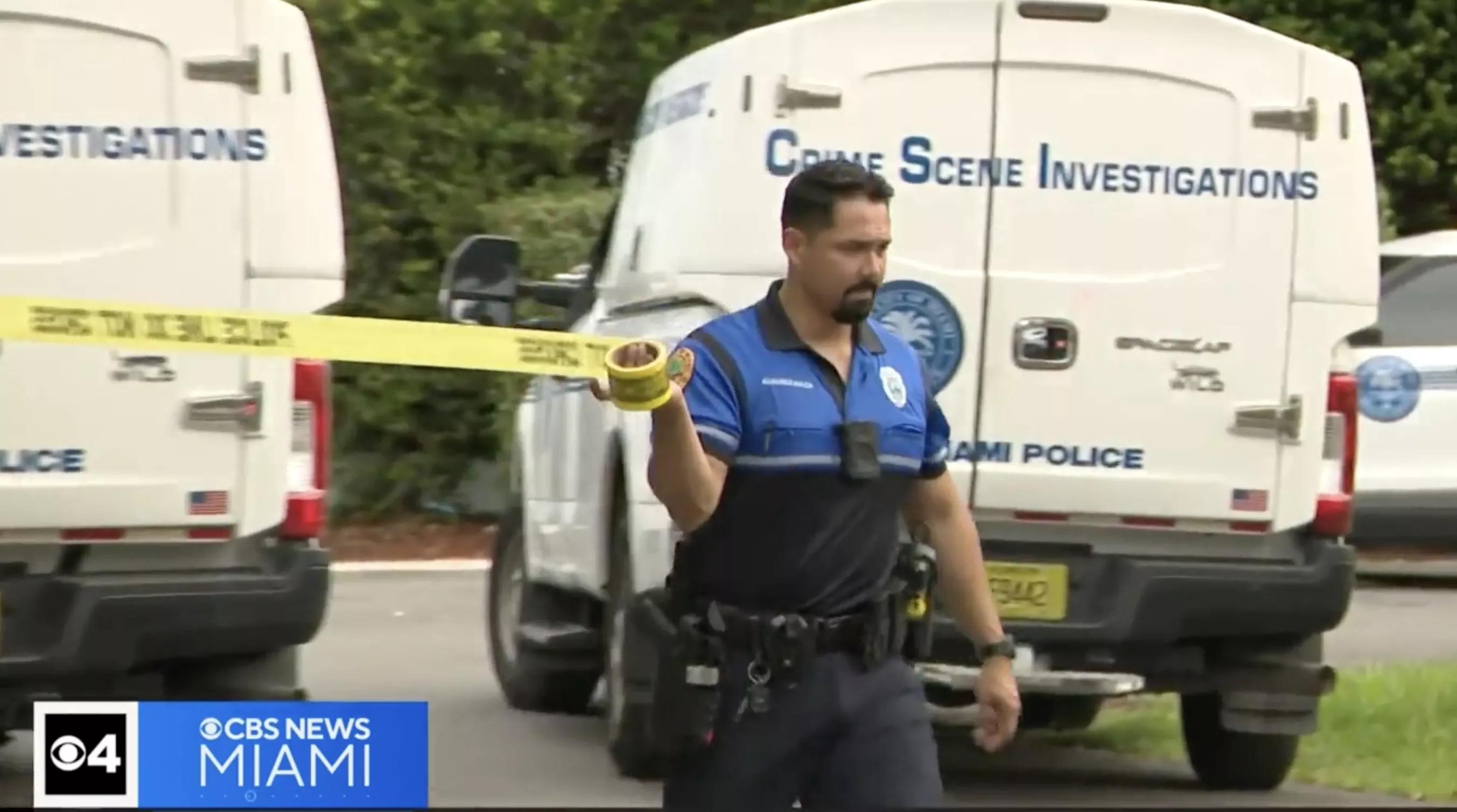 a Miami law enforcement officer pulls crime scene tape across a residential street with police crime scene investigation vehicles in the background