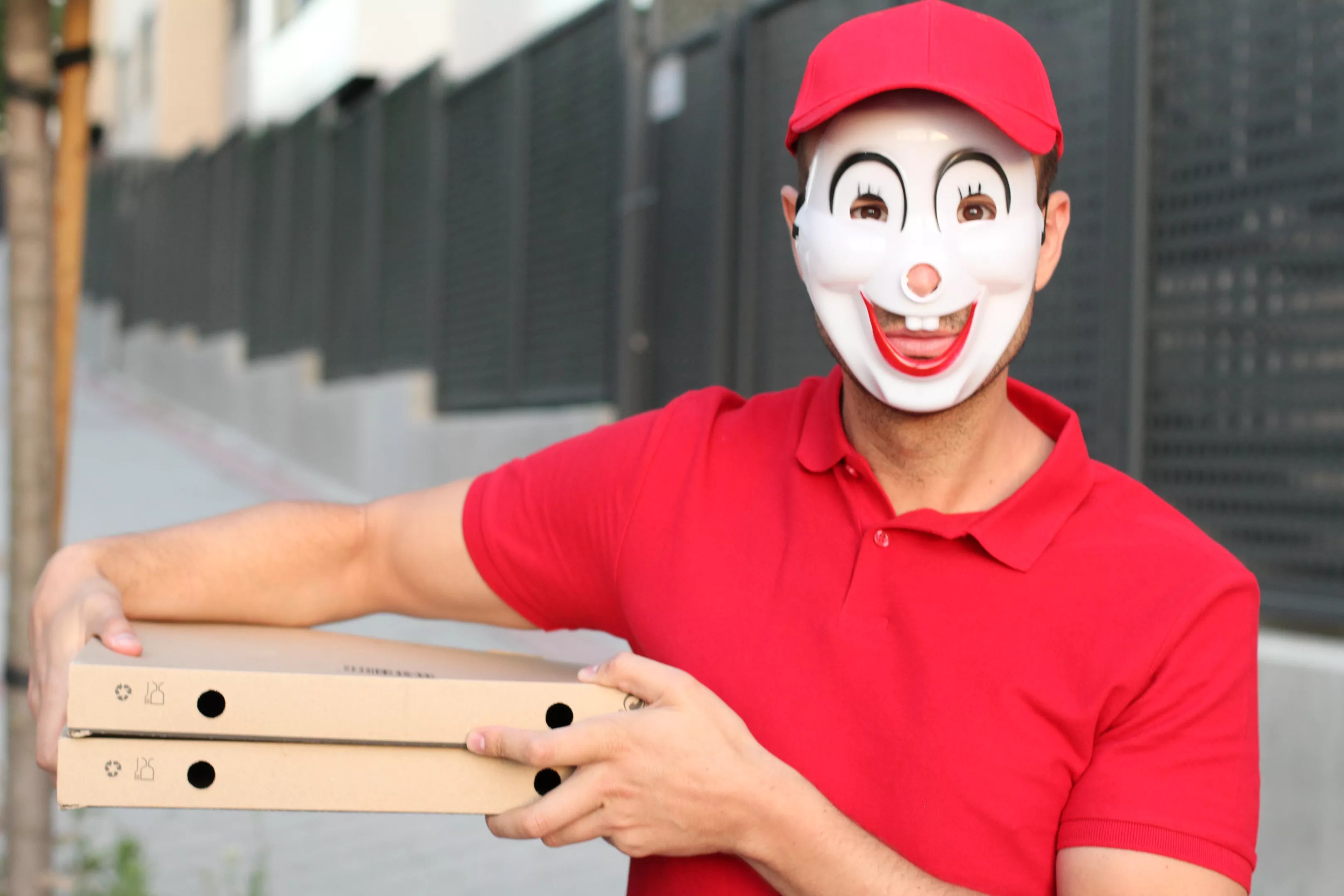 A man wearing a strange, doll-like mask holds a few pizza boxes at his side