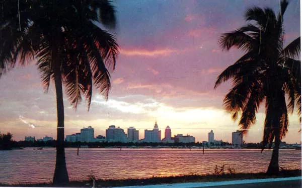 color postcard from 1956 featuring a photo of the Miami skyline, flanked by two palm trees, with Biscayne Bay in the middle distance