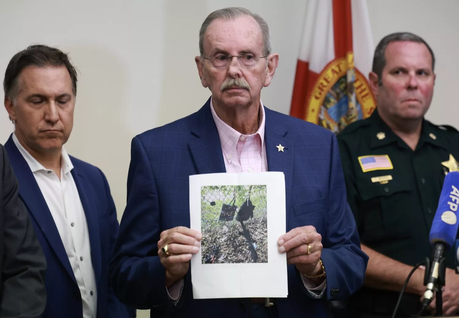 photo of a gray-haired, mustachioed man wearing wire-rimmed glasses looking somber as he holds up a color printout showing a rifle and other items near a chain-link fence