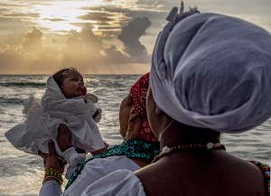 A photo by Woosler Delisfort depicting a spiritual baptism ritual with a child held at the ocean’s edge during sunrise