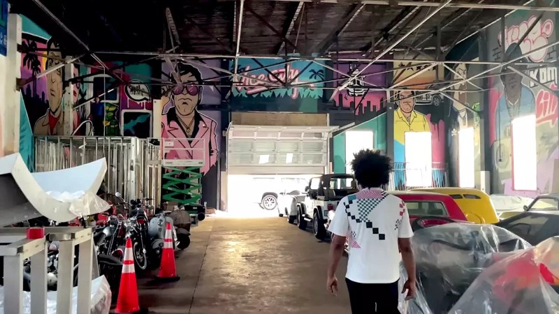 Elo King walks through the interior of Vice City District, surrounded by colorful Miami Vice-inspired murals, vintage cars, and motorcycles in a large industrial warehouse space.