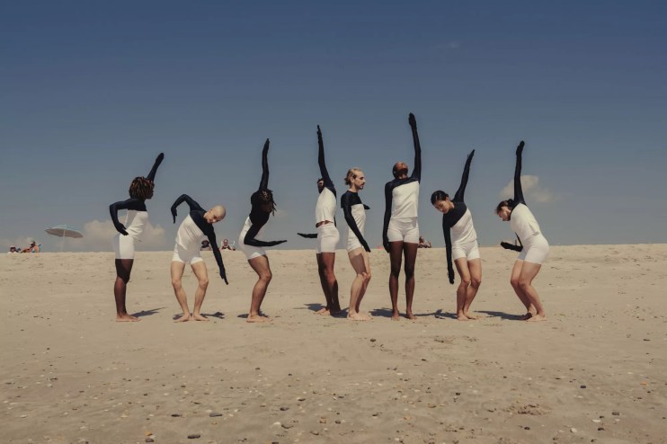 dancers in pose on a beach in white shirts and shorts with arms and hands covered in black cloth