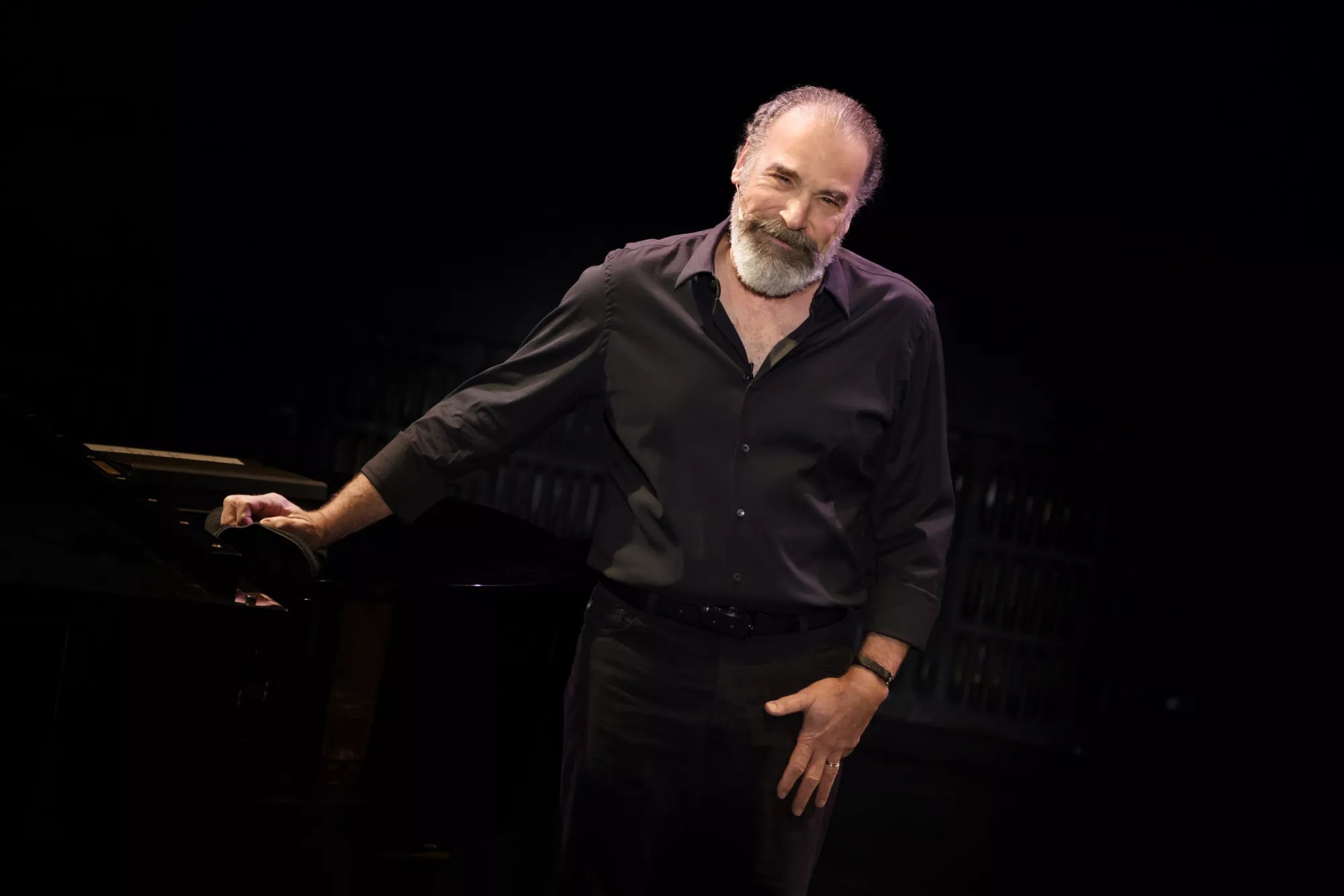 portrait of Mandy Patinkin in a black button-down shirt and pants, leaning on a black piano inside a black box theater