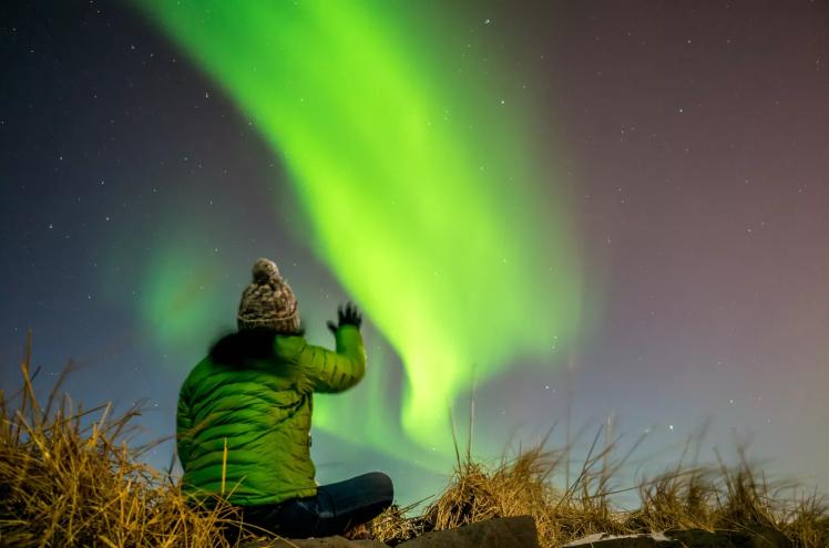 photo of a person in a beanie and thick green jacket looking up at green Northern Lights