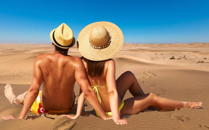 photo-illustration depicting a bathing suit clad couple avec straw hats appearing to sunbathe in a Gambian desert as opposed to on tropical a beach