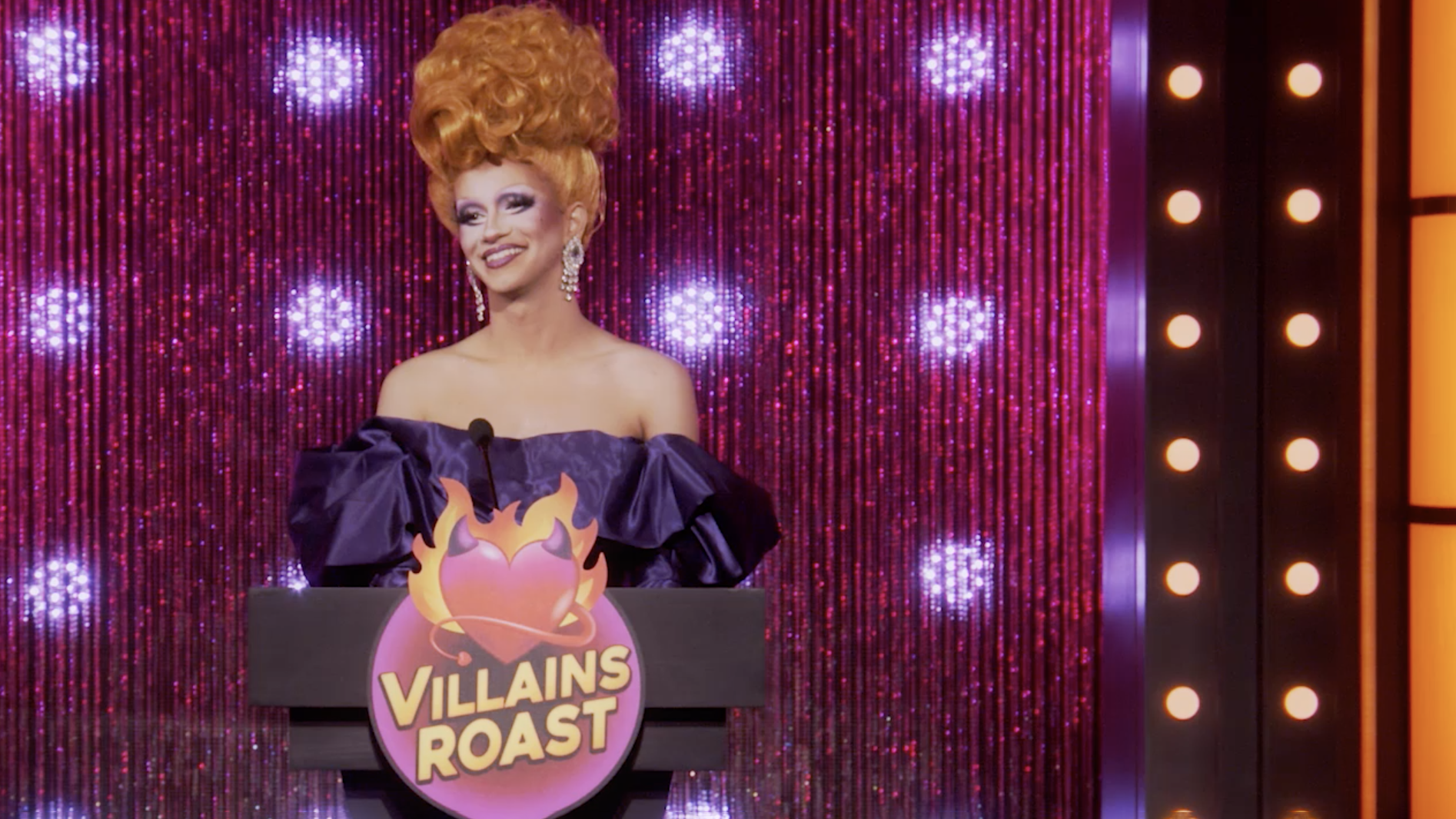 Drag queen Suzie Toot stands in front of a podium with a sign reading Villains Roast
