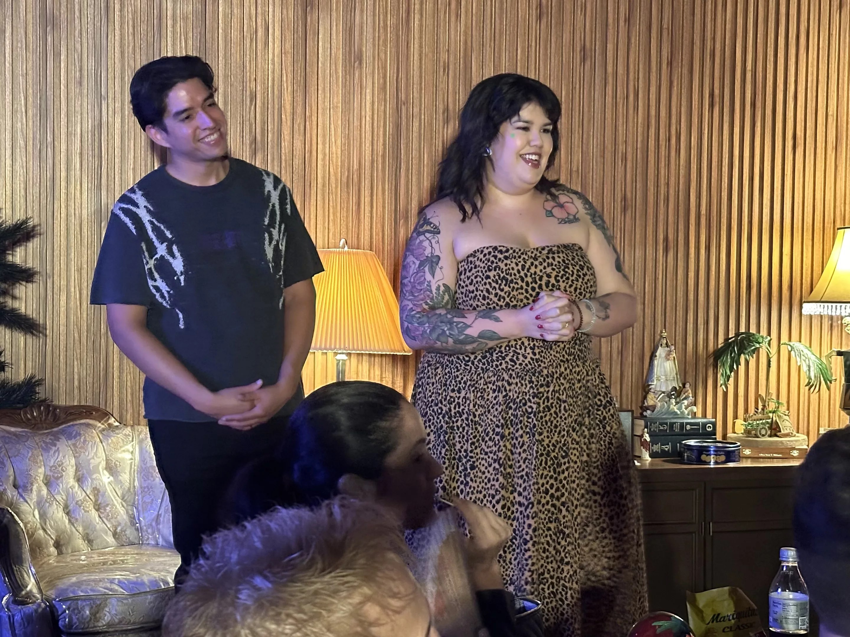 a performer speaks to an audience in a living room as another performer stands next to her