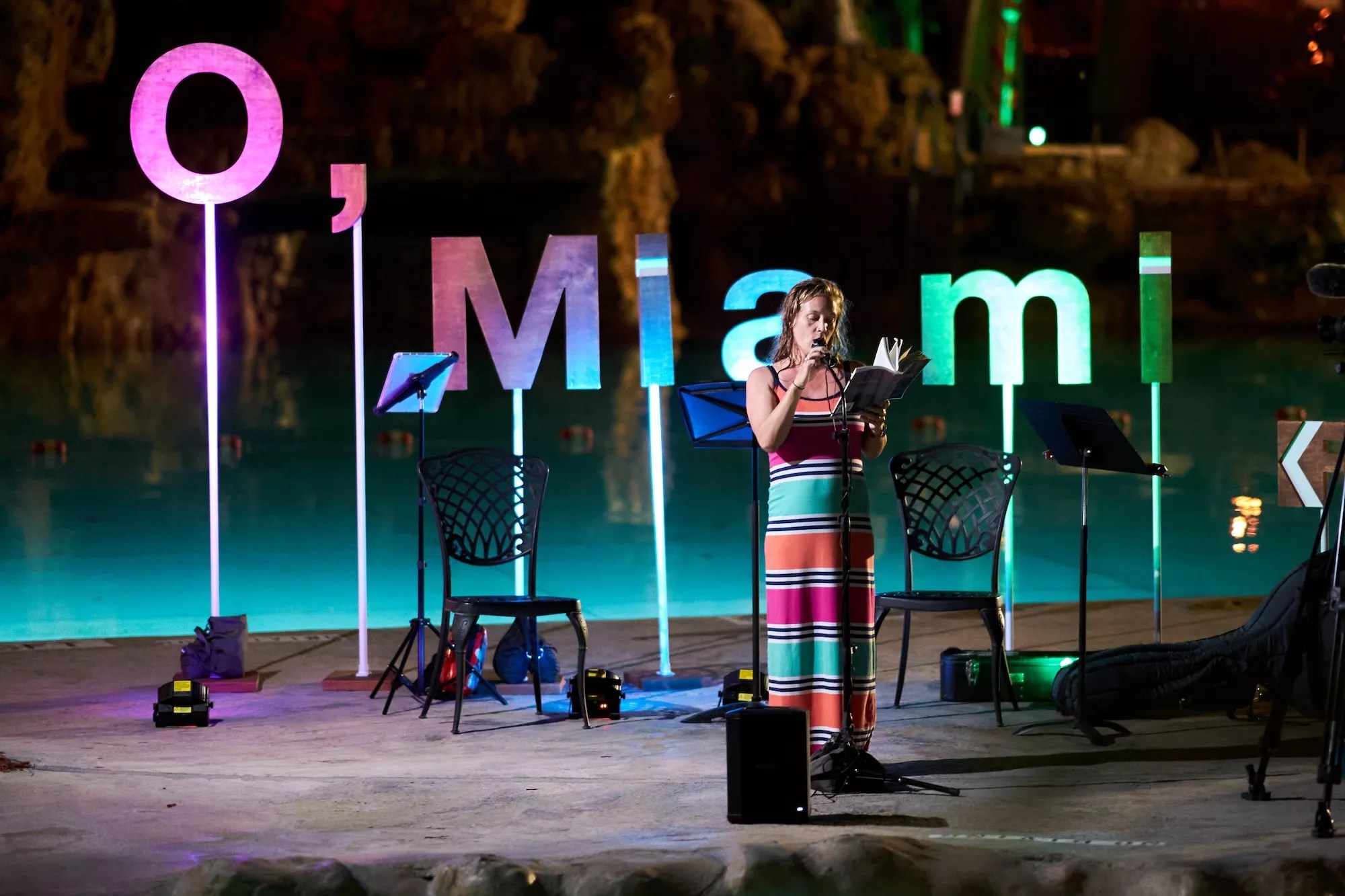 a person reads a poem in front of a backdrop that reads, "O, Miami"