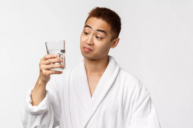 Portrait of Asian man in bathrobe looking skeptical while holding glass of water