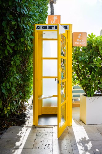 yellow old-school telephone booth. a sign on the booth reads "astrology hotline." another sign protruding from its side reads "what's your sign"