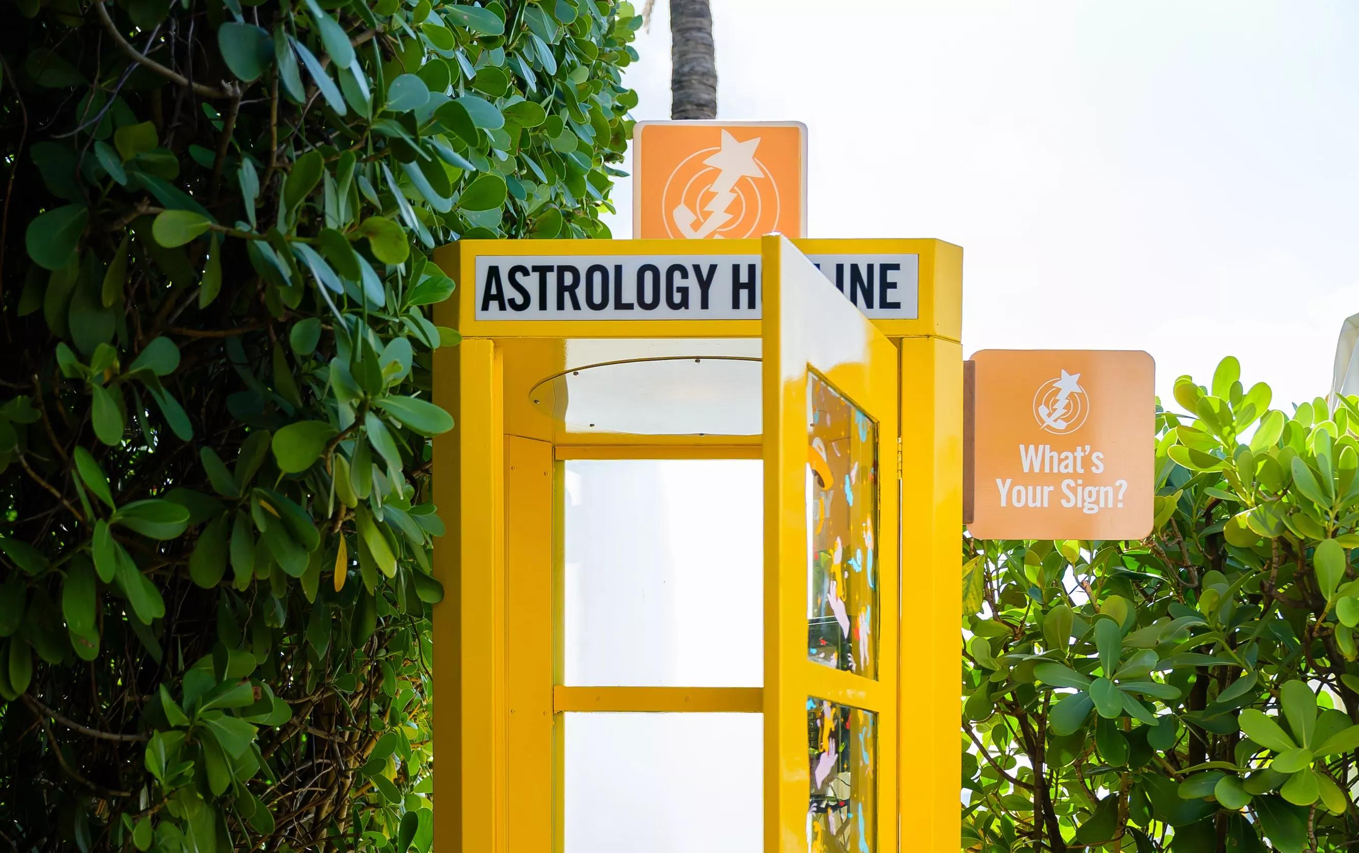 yellow old-school telephone booth. a sign on the booth reads "astrology hotline." another sign protruding from its side reads "what's your sign"