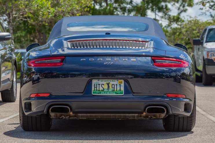Rear end of a dark blue convertible Porsche 911 in a parking lot with a golfer on the license plate and dual exhausts