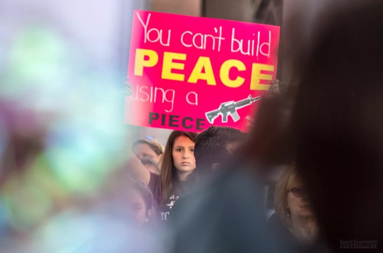 A girl holds a poster that reads "You can't build peace using a piece" with a photo of a gun.
