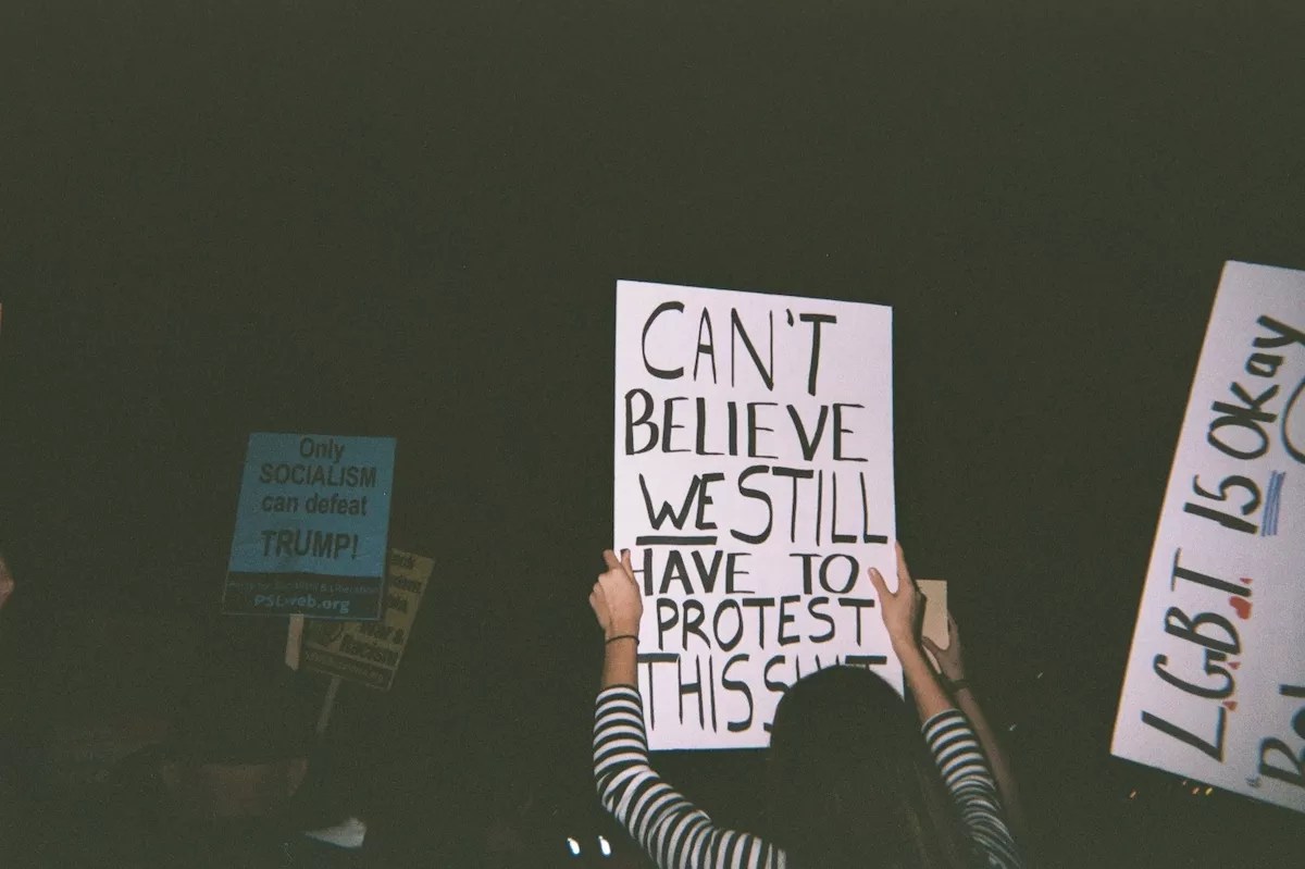 Someone holds a sign that reads, "CAN'T BELIEVE WE STILL HAVE TO PROTEST THIS SHIT."